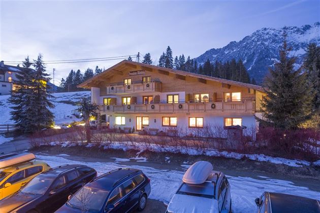 Ein gemütliches Holzgebäude in den Alpen, umgeben von schneebedeckten Bergen und Tannenbäumen. Im Vordergrund sind verschiedene Autos geparkt.