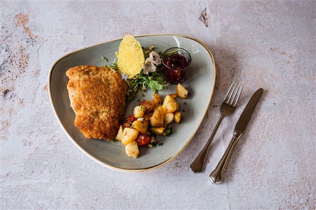 Ein gut angerichtetes Gericht mit paniertem Fisch, Beilagen aus Kartoffeln und Gemüse sowie einer kleinen Sauce. Das Essen ist auf einem eleganten Teller serviert.
