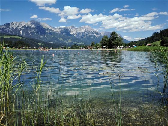 Ein klarer See umgeben von Wiesen und Bergen. Der Himmel ist blau und mit einigen Wolken versehen.