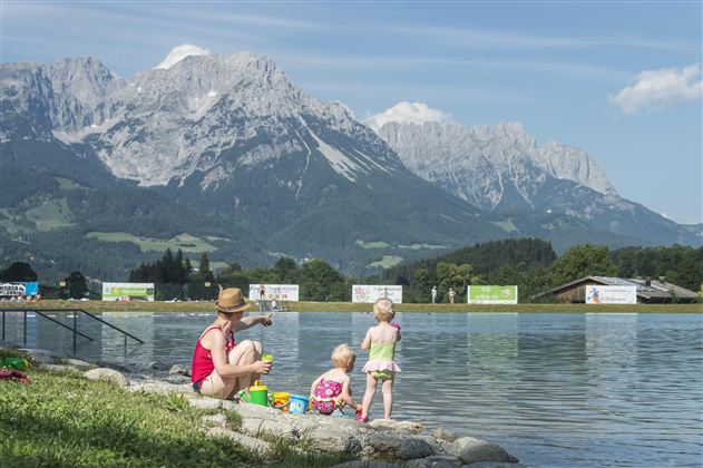 Eine Mutter und zwei kleine Kinder sitzen am Ufer eines Sees mit Blick auf die Berge. Der Himmel ist klar und sonnig, ideal für einen Familientag im Freien.