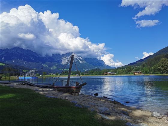 Ein ruhiger See umgeben von Bergen und grünen Wiesen. Die Wolken sind groß und weiß vor einem klaren blauen Himmel.