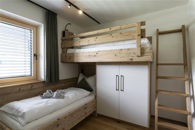 A modern bedroom with a wooden bunk bed and a single bed. The walls are light and there is a large window with a view of the surroundings.