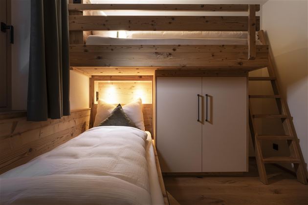 A cozy bedroom with a loft bed and a single bed side. The room is made of wood and features a modern wardrobe.