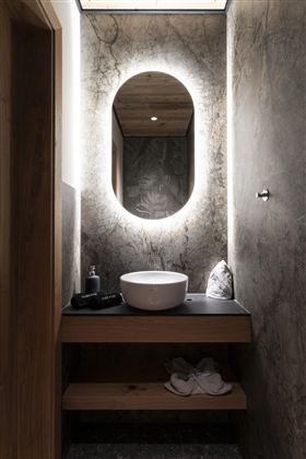 A modern bathroom with elegant lighting and a round mirror. A minimalist wooden vanity top with a white basin and fluffy slippers underneath.