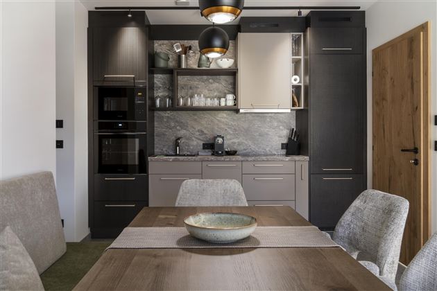 A modern kitchen with dark cabinets and white accents. A dining area with a wooden tray and comfortable chairs is also visible.