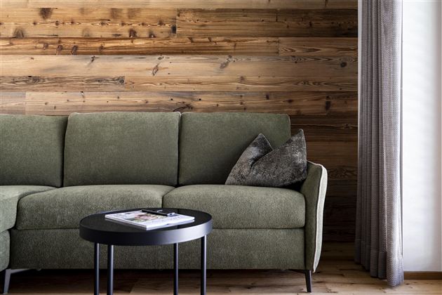 A modern living room with a comfortable, gray-green sofa and a small black table. The wooden wall combined with curtains creates a cozy atmosphere.