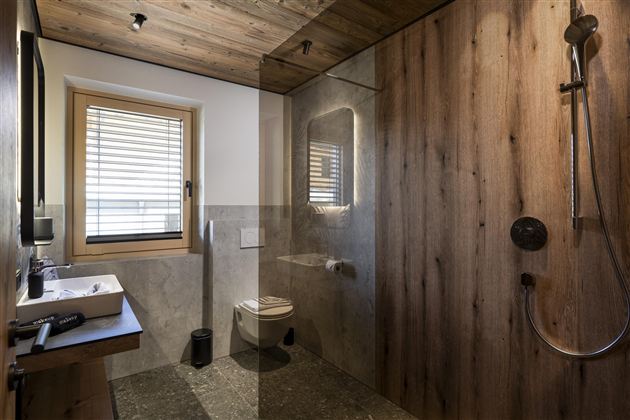 A modern bathroom with wooden paneling and gray tiles. It includes a glass shower, a sink, and a small window.