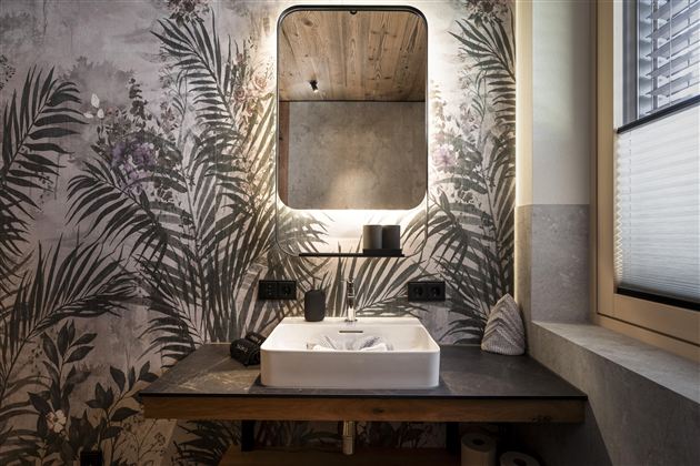 A modern bathroom with a large mirror and a sink on a wooden table.  
Walls with a tropical plant motif create a relaxed atmosphere.