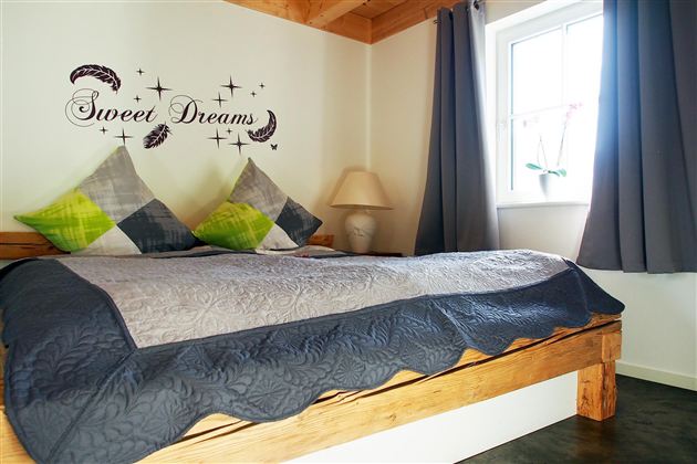 A cozy bedroom with a wooden bed and decorative pillows. On the wall, there is the inscription "Sweet Dreams," and there is a window with curtains.