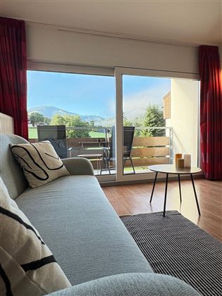 Een gezellige woonkamer met een lichte bank en uitzicht op de bergen. Buiten is er een balkon met tuinmeubelen en een mooi uitzicht.