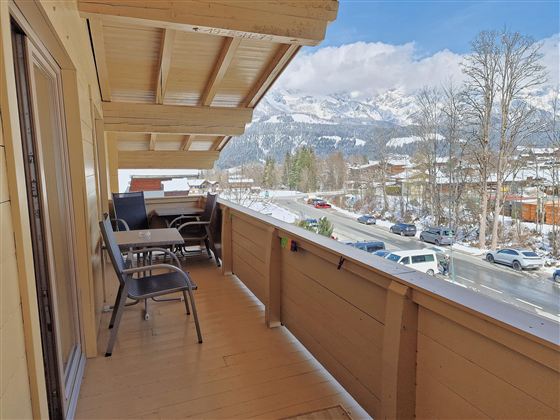 A balcony with wooden cladding, on which there are chairs and a table. In the background, snow-covered mountains and a clear blue sky can be seen.
