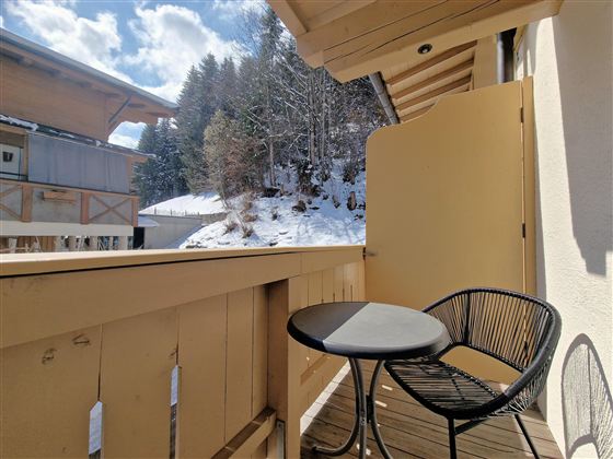 A balcony with a small table and a chair, surrounded by snow-covered trees. The sky is clear and blue, providing a beautiful view.