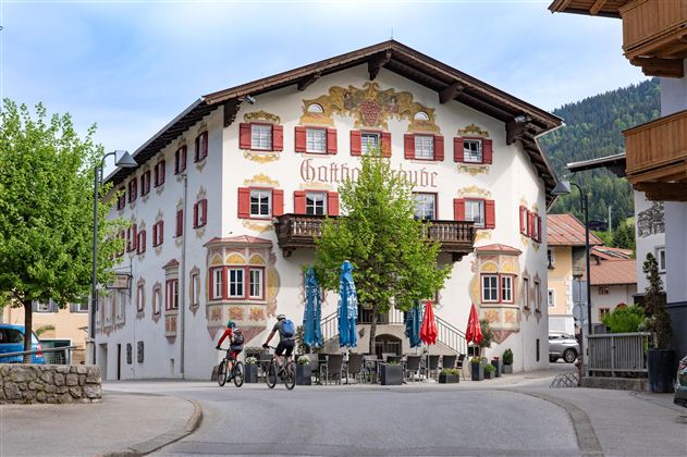 A historic building with colorful windows and a balcony. Two cyclists ride past a shady tree.