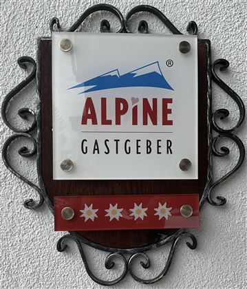 Ein Logo mit dem Schriftzug "ALPINE GASTGEBER" und stilisierten Bergen. Die Farben sind rot, blau und weiß, mit Edelweiß-Blumen am unteren Rand.