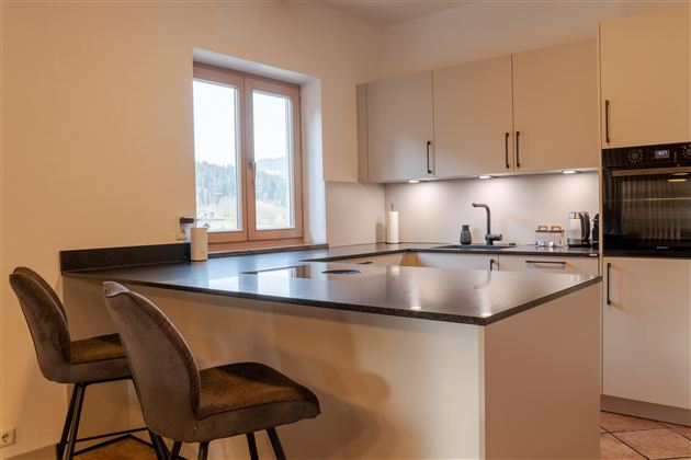 Een moderne keuken met een centraal kookeiland en hoge stoelen. Veel daglicht valt door het raam en zorgt voor een lichte sfeer.