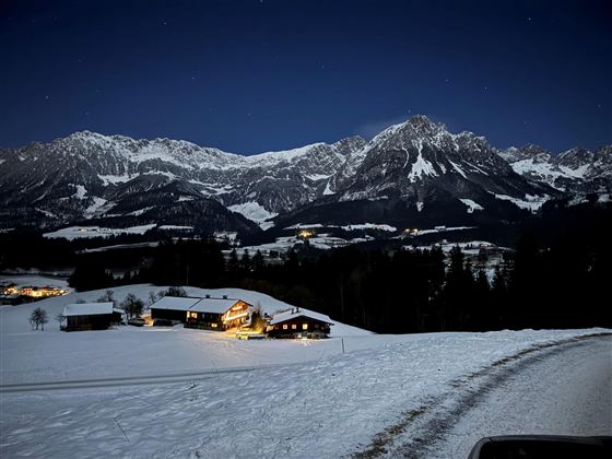 Eine verschneite Landschaft bei Nacht mit beleuchteten Häusern. Im Hintergrund erheben sich beeindruckende Berge unter einem klaren Sternenhimmel.