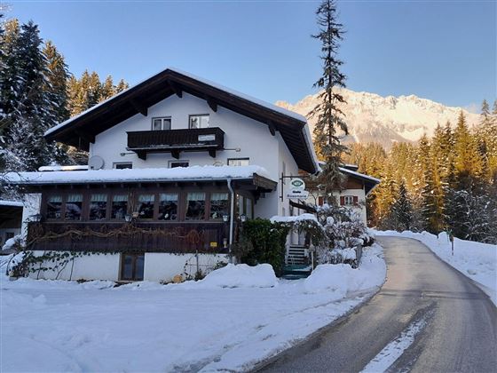Ein gemütliches Haus in einer winterlichen Landschaft, umgeben von Schnee und Tannenbäumen. Im Hintergrund sind majestätische Berge sichtbar.