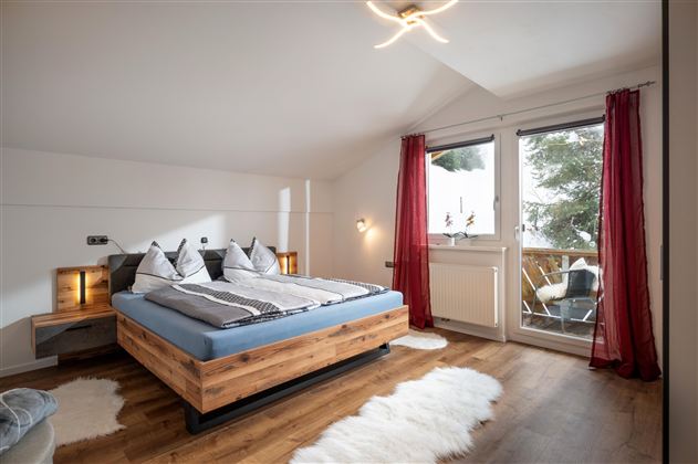 Ein gemütliches Schlafzimmer mit einem großen Doppelbett und modernen Holzdetails. Große Fenster lassen viel Tageslicht herein und bieten Zugang zu einem Balkon.