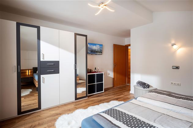 Ein modernes Schlafzimmer mit einem großen Spiegelkleiderschrank und einem gemütlichen Bett. Die Wände sind hell und der Boden ist aus Holz.