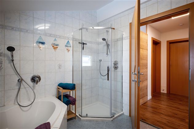 Ein modernes Badezimmer mit einer Duschkabine und einer Badewanne. Der Boden ist aus Holz, und es gibt Zugang zu einem weiteren Raum.