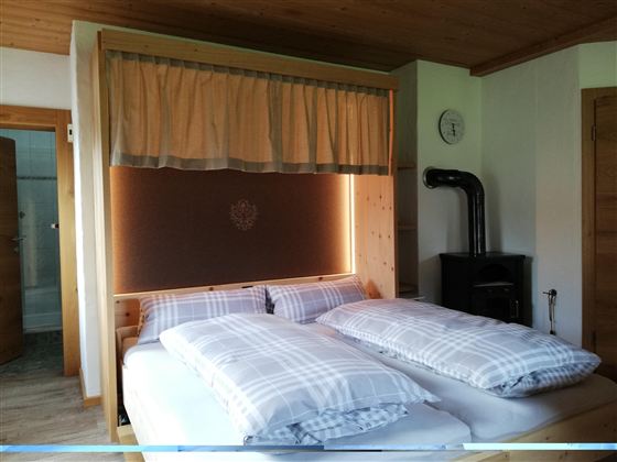 Ein gemütliches Schlafzimmer mit einem großen Bett und karierten Bettdecken. Die Wände sind aus Holz und es gibt einen kleinen Ofen.