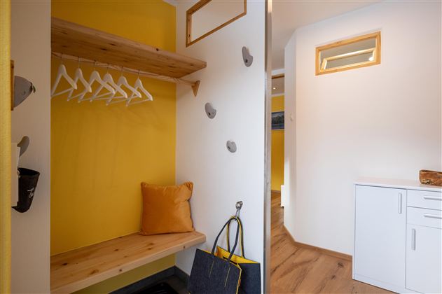 Een heldere entree met een houten bank en gele muren. Aan de muur hangen kledinghaken en er is een kleine commode.