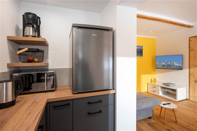 Een moderne keuken met een koelkast, een magnetron en een koffiezetapparaat. De ruimte heeft een lichte houten afwerking en een open overgang naar een woonruimte met een televisie.