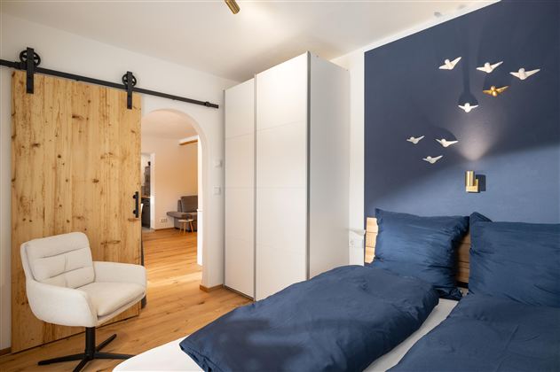 Een moderne slaapkamer met een blauwe accentwand en een witte kast. Een comfortabele stoel staat naast een bed met blauwe beddengoed.