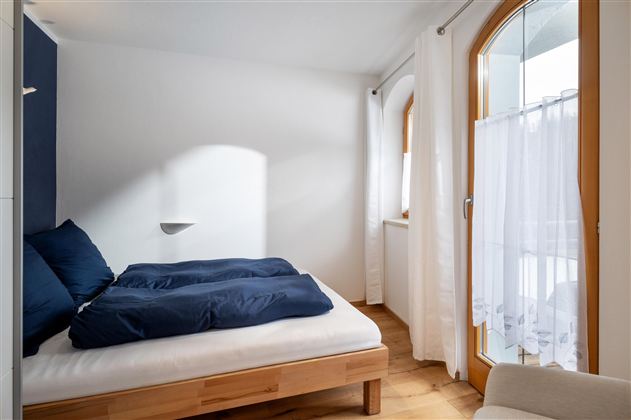 Een knusse slaapkamer met een bed en blauwe kussens. Grote ramen zorgen voor veel licht en uitzicht naar buiten.