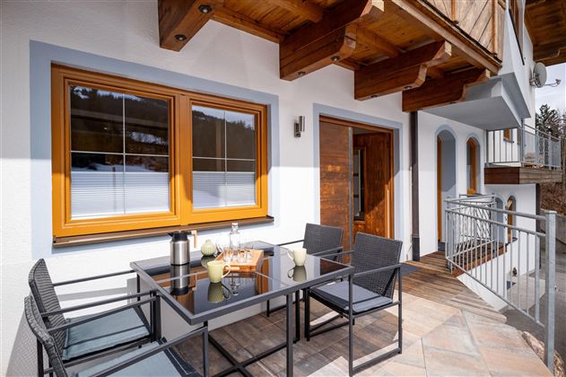 Een gezellige veranda met een tafel en stoelen. Op de achtergrond zijn grote ramen en een houten deur zichtbaar.