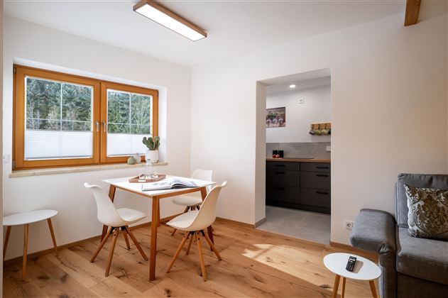 Een gezellige woonkamer met een houten tafel en moderne stoelen. De keuken is op de achtergrond zichtbaar, en grote ramen laten veel licht binnen.