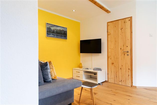 Een gezellige woonkamer met een gele muur en een moderne televisie. Er is een bank, een kleine tafel en een houten deur.