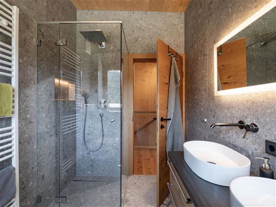 A modern bathroom with a glass shower and sink. The walls are in neutral gray tones, and wooden details create a warm atmosphere.