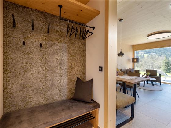 A modern entrance area with a bench and coat hooks. In the background, a cozy living area with a table and chairs is visible.