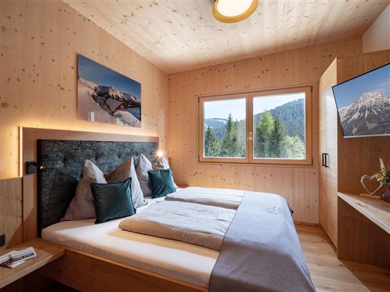 A modern bedroom with bright wood decor and large windows. The room is cozily furnished and offers a view of the surrounding mountains.