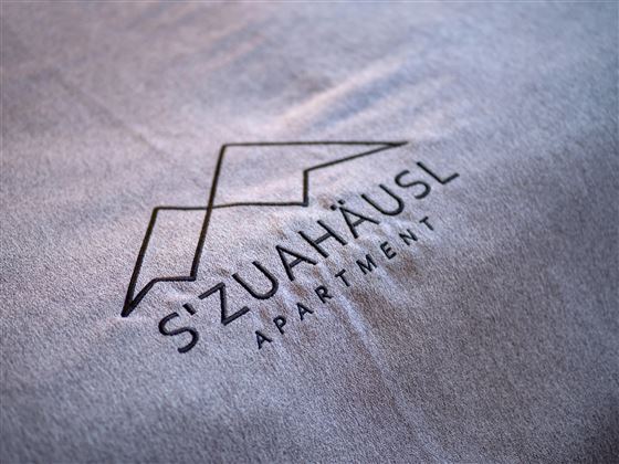 A modern logo for the apartment "S'ZuaHäusl". The design is simple and elegant on a gray background.
