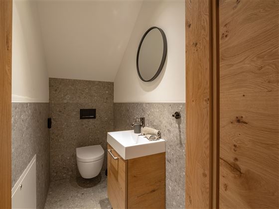 A modern bathroom with a wall-mounted toilet and a stylish wooden vanity combination. The space has a bright and minimalist design with a round mirror.