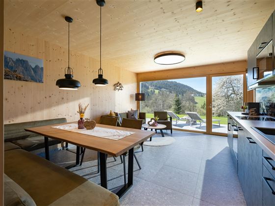 A modern kitchen-living room with wooden walls and large windows. The space offers a view of the greenery and is cozily furnished.