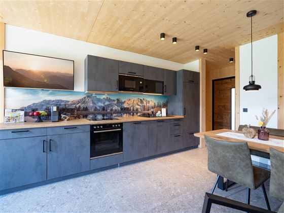 A modern kitchen with gray cabinets and a wooden ceiling. A dining table with chairs is next to the kitchen unit.