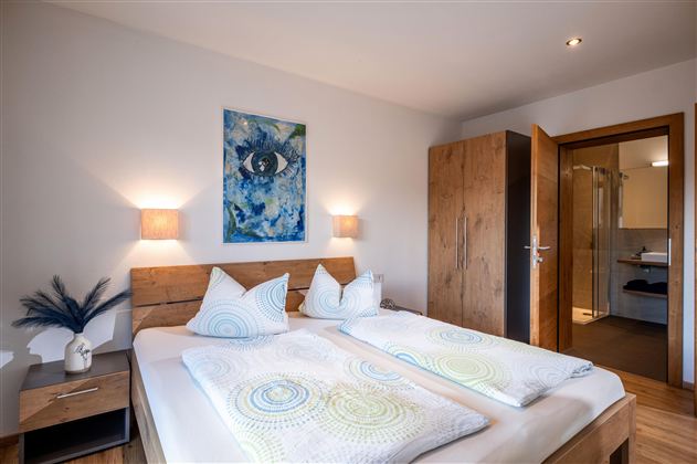 A modern bedroom with a double bed and two nightstands. A painting hangs on the wall, and there is access to the bathroom.