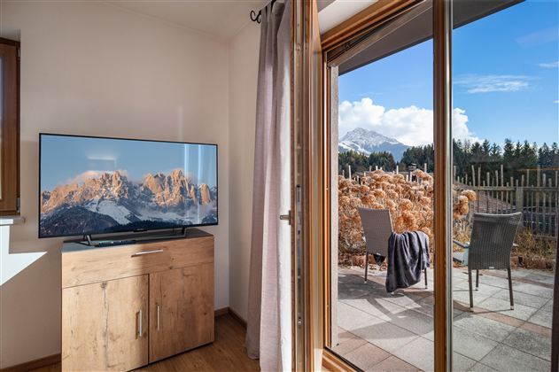 A cozy room with a television and a view of the mountains. Outside is a terrace with chairs and a beautiful landscape.