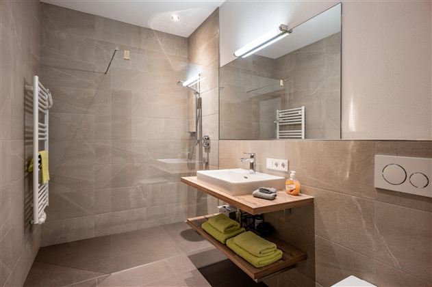 A modern bathroom with a large shower and a bright sink. The walls are painted in neutral tones and there are towels on a wooden shelf.