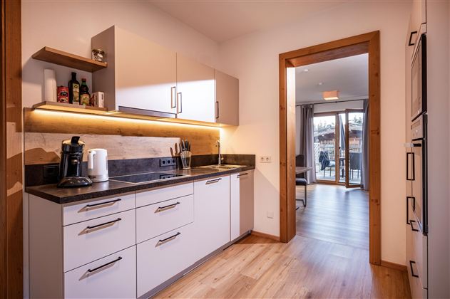 A modern kitchen with a wooden floor and bright lighting. Through the open door, a living area with access to the balcony is visible.