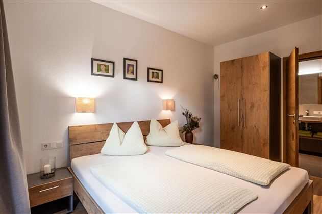 A cozy bedroom with a double bed and soft lighting. Some pictures hang on the wall, and a wardrobe stands in the room.