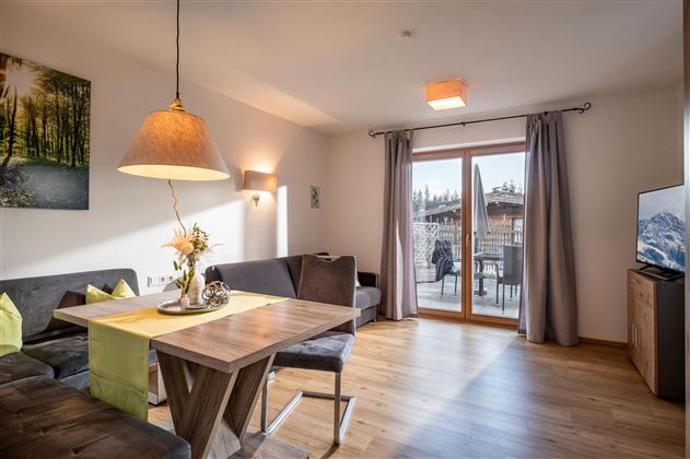 A modern living room with a dining table and a cozy couch. Large windows provide a view of the terrace outside.