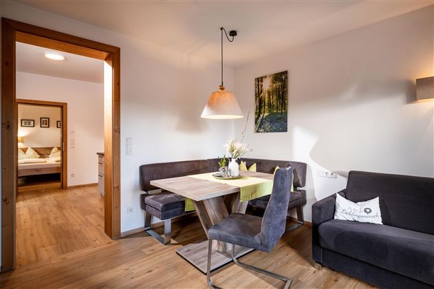 A cozy living area with a dining table, chairs, and a sofa corner. A picture hangs on the wall, and the light is pleasant and warm.