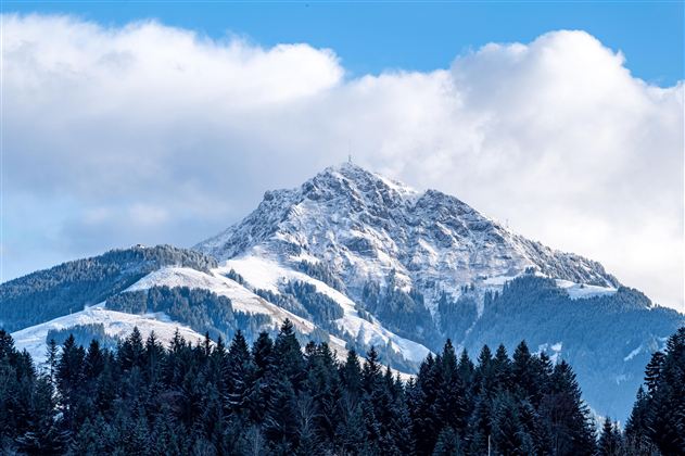 A majestic mountain with a snow-capped peak and green forests in the foreground. The sky is clear with a few clouds.