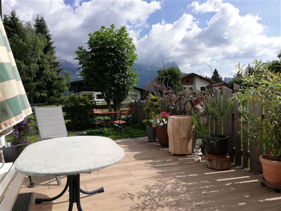 Eine Terrasse mit einem Tisch und einem Stuhl, umgeben von Pflanzen und Bäumen. Der Himmel ist teilweise bewölkt mit etwas Sonnenschein.