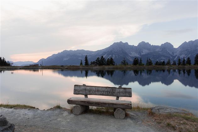 Een houten bank staat aan de oever van een rustig meer, omgeven door bergen. De lucht heeft zachte kleuren bij zonsondergang.