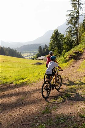 Twee kinderen fietsen op een smal pad in een pittoresk landschap. Op de achtergrond zijn bergen en bossen te zien.